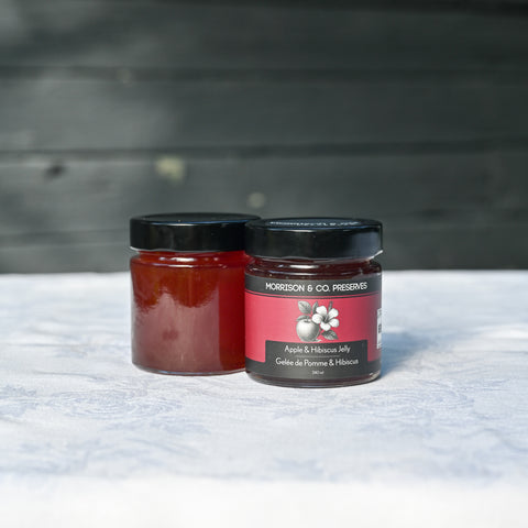 Two glass jars of Morison & Co Preserves Apple and Hibiscus Jelly, displayed on a white patterned surface against a dark wooden background.