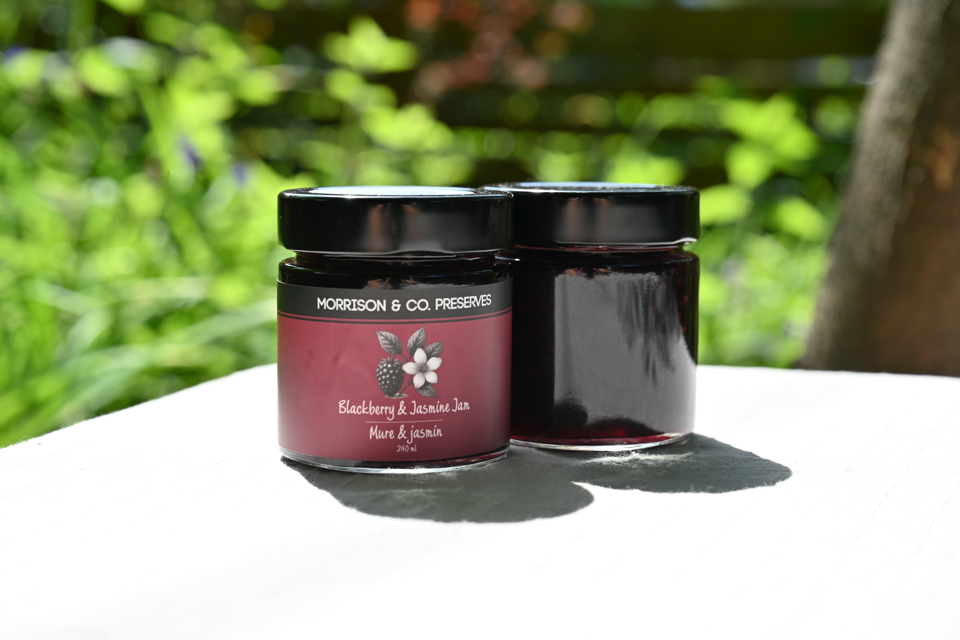 Two jars of blackberry and jasmine tea jam with a label on a white surface outdoors