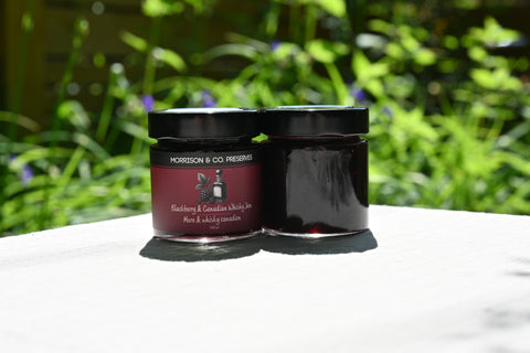Two jars of blackberry and Canadian whisky jam on a white surface with a blurred green background.