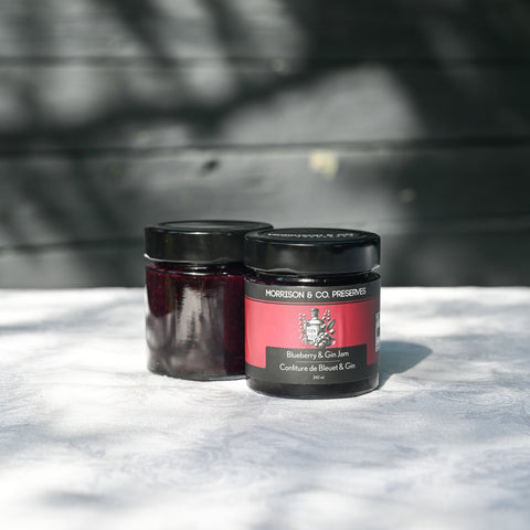 Two glass jars of preserves with black lids, one in the foreground labeled 'Morrison & Co Preserves Blueberry & Gin Jam' and the other behind it, set on a white textured surface with a dark wooden background.