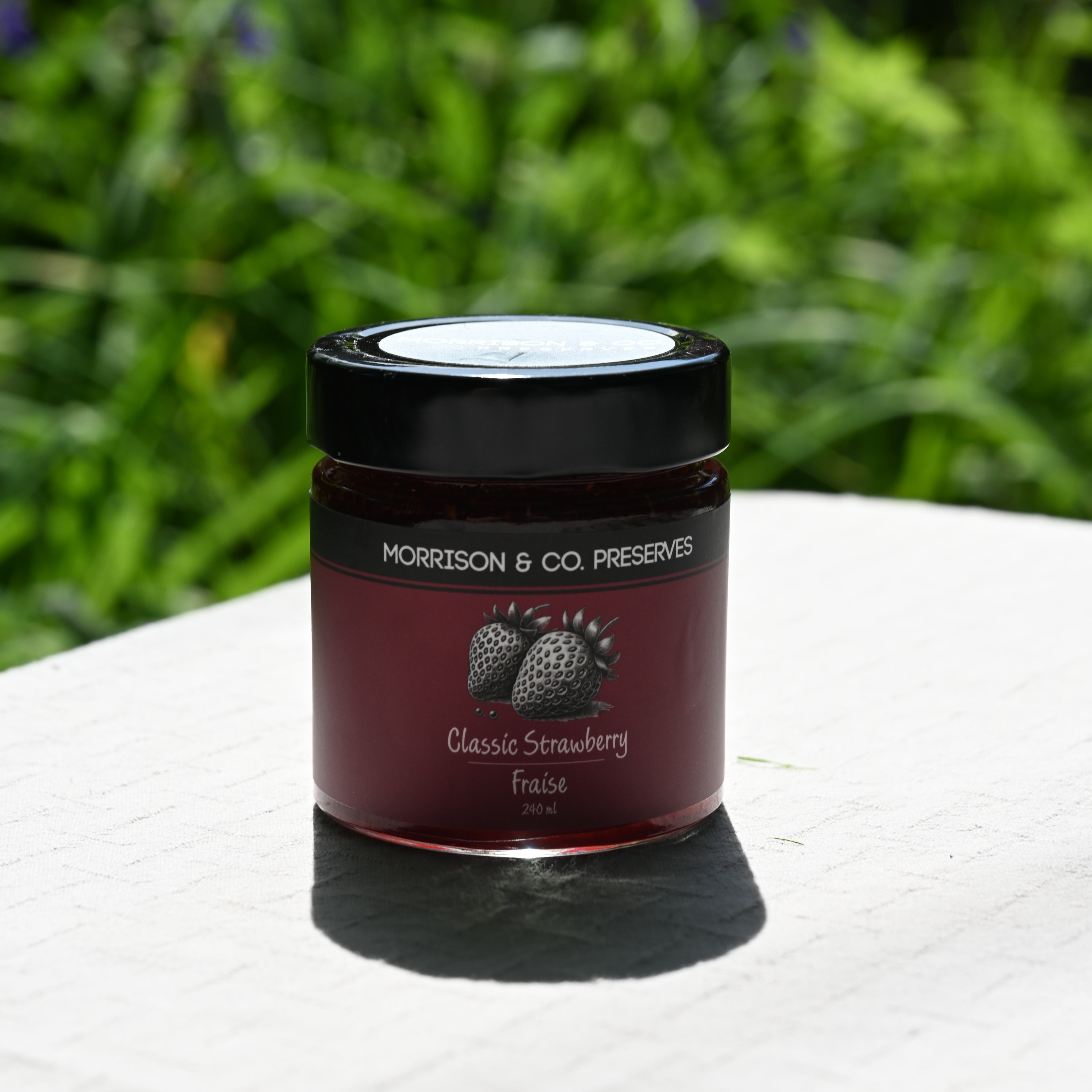 Jar of Morrison & Co. Preserves Classic Strawberry on a white surface with greenery in the background