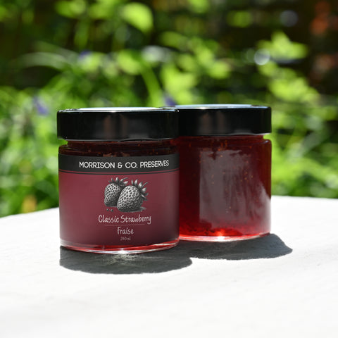 Two jars of Morrison & Co. Preserves Classic Strawberry Fraise on a white surface with a green blurred background.