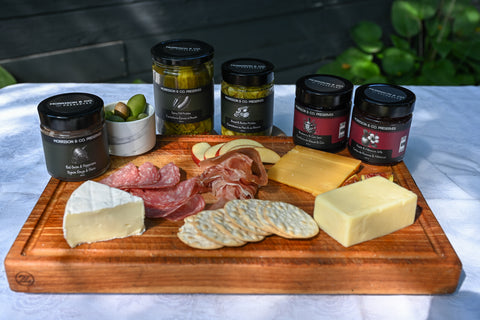 A wooden charcuterie board arranged with cheeses, cured meats, crackers, and several jars of Morrison & Co. preserves, set on a white tablecloth with a dark wooden wall and greenery in the background.