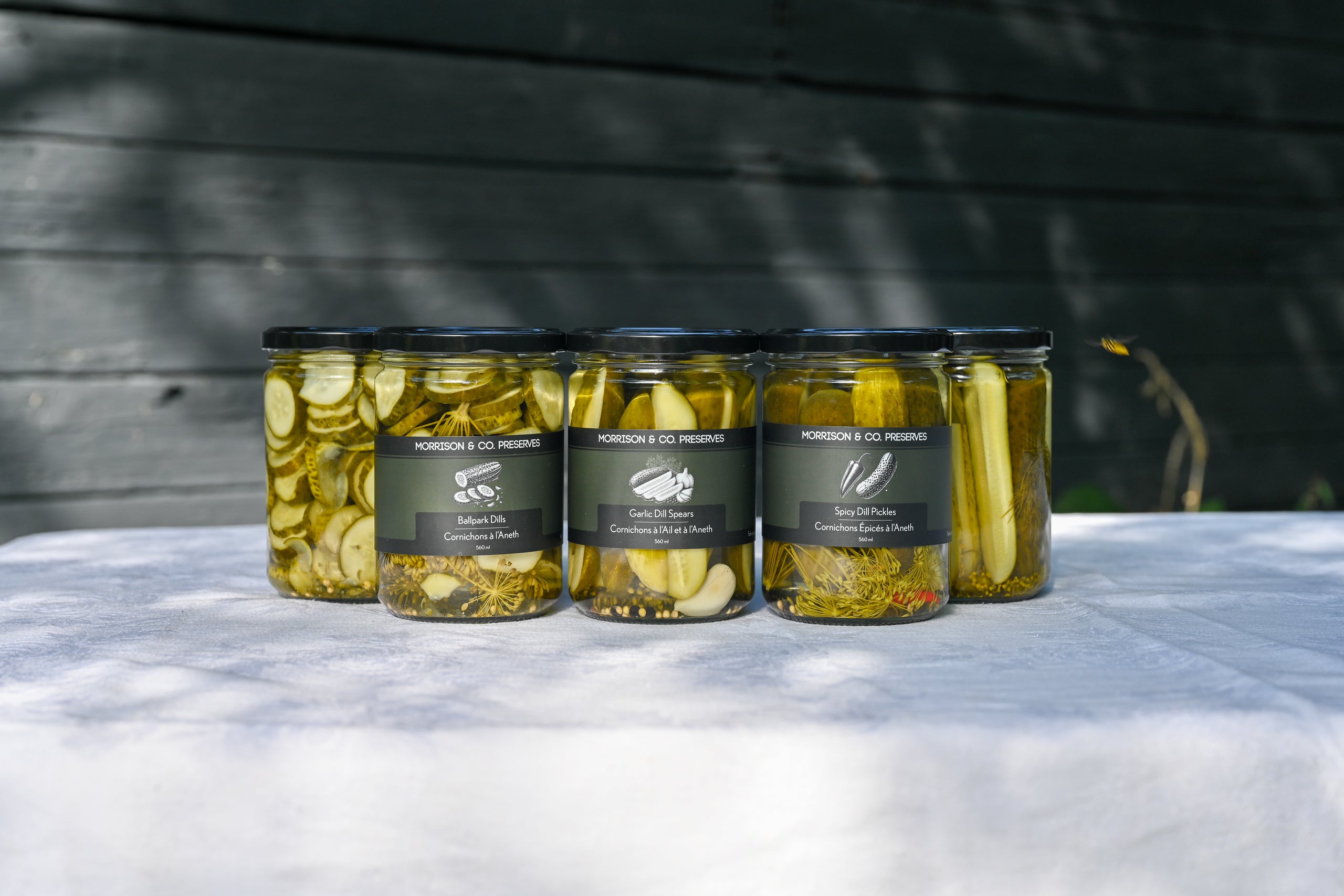 Five glass jars of pickles arranged in a row on a white tablecloth, with a dark wooden wall in the background. The jars have black lids and dark labels with white text