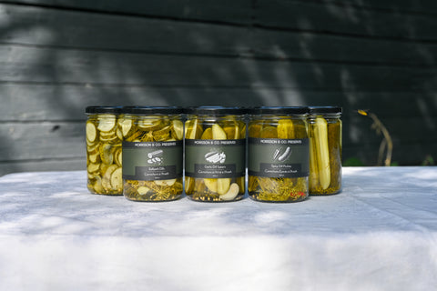 Five glass jars of pickles arranged in a row on a white tablecloth, with a dark wooden wall in the background. The jars have black lids and dark labels with white text