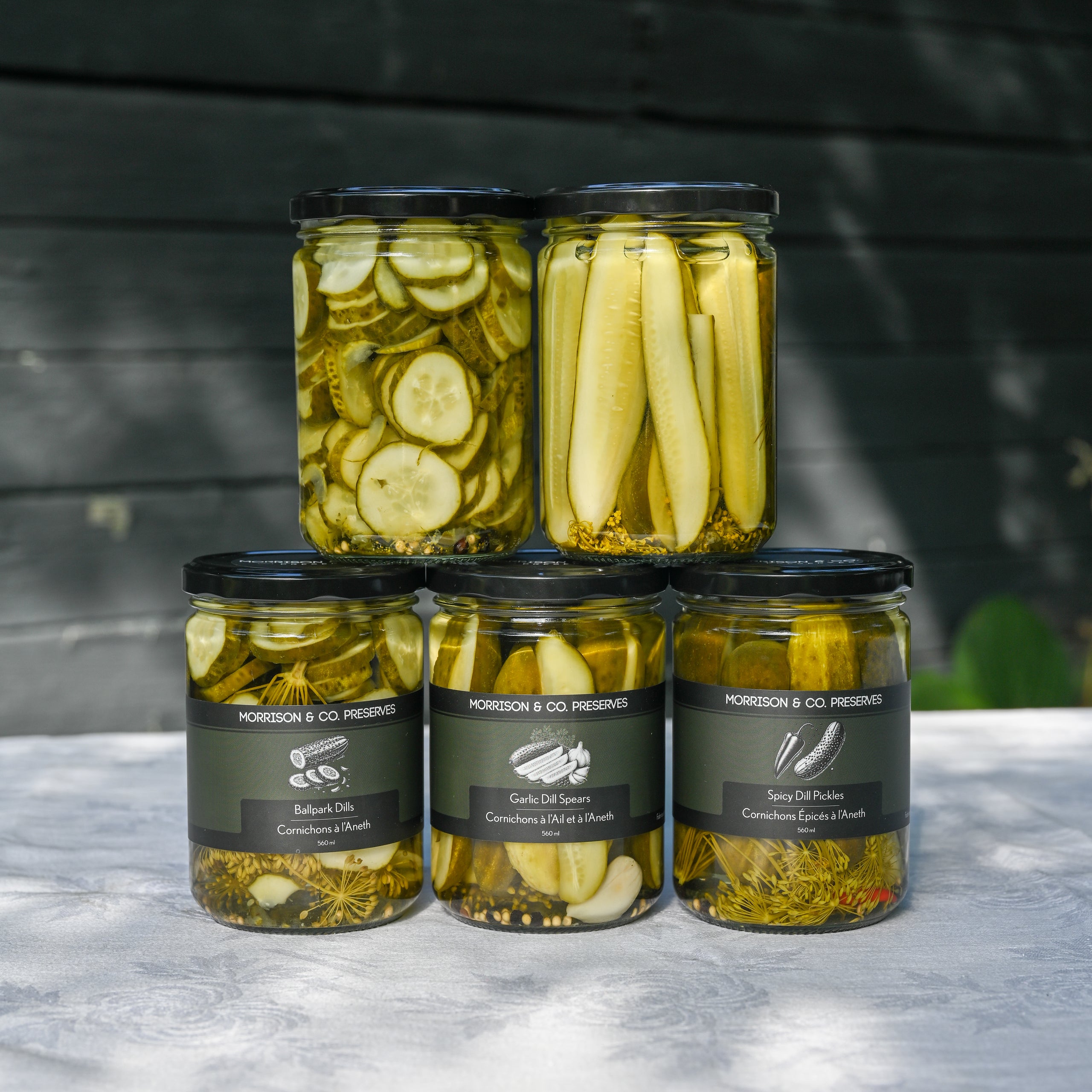 Four glass jars of Morrison & Co Preserves pickles arranged in a pyramid on a white tablecloth against a dark wood background. The top row contains two jars with sliced cucumbers and pickles, and the bottom row contains three different types of dill pickles. 