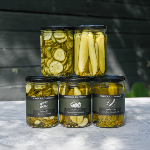 Four glass jars of Morrison & Co Preserves pickles arranged in a pyramid on a white tablecloth against a dark wood background. The top row contains two jars with sliced cucumbers and pickles, and the bottom row contains three different types of dill pickles. 
