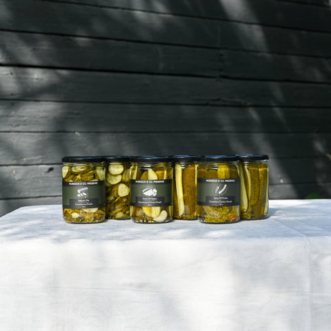 Five glass jars of Morrison & Co Preserves dill pickled cucumbers with black lids and black labels, arranged on a white draped tablecloth against a dark wooden wall with leaf shadows.