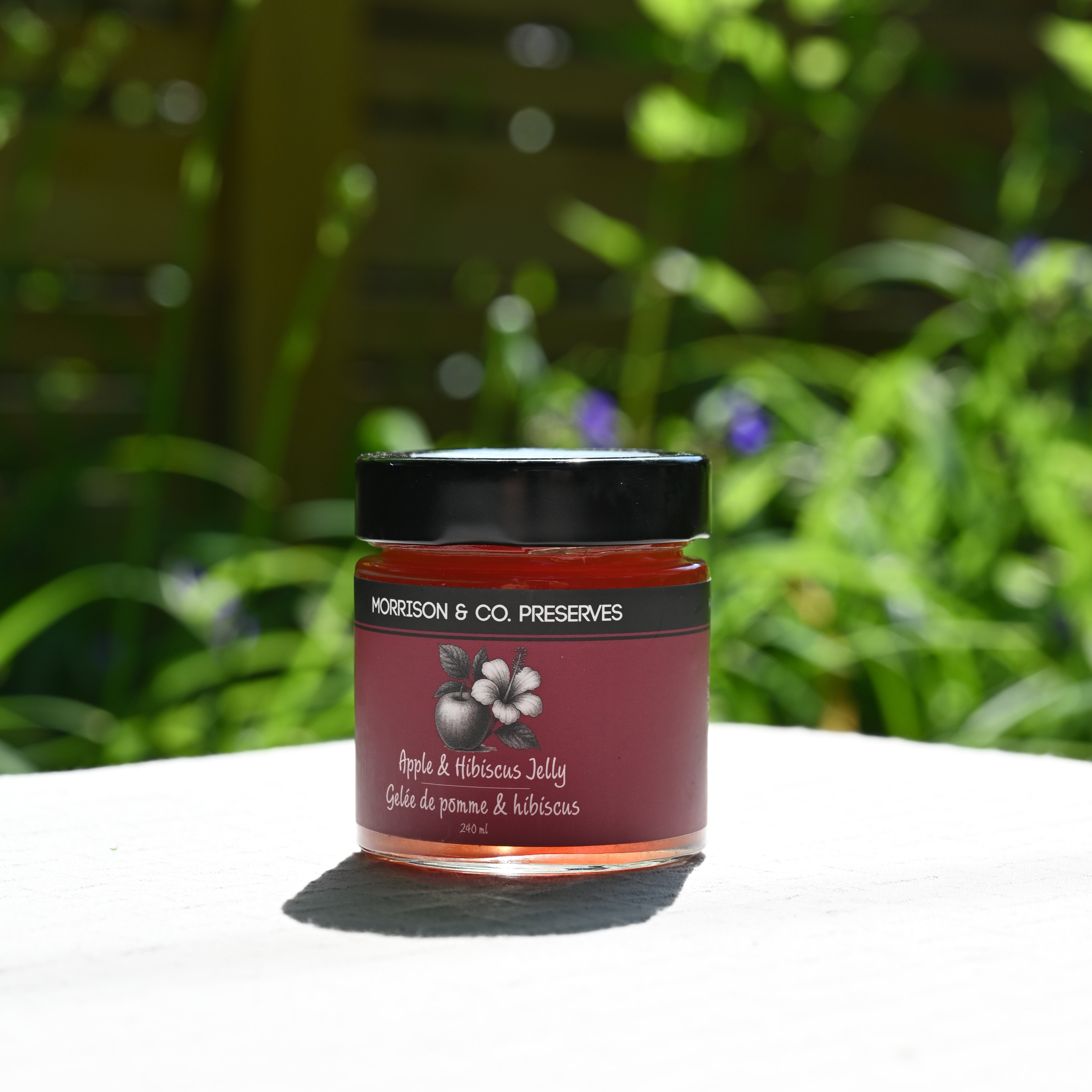 Jar of apple and hibiscus tea jelly with a black lid on a white surface, blurred greenery in the background