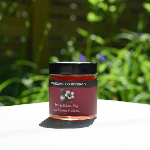 Jar of apple and hibiscus tea jelly with a black lid on a white surface, blurred greenery in the background