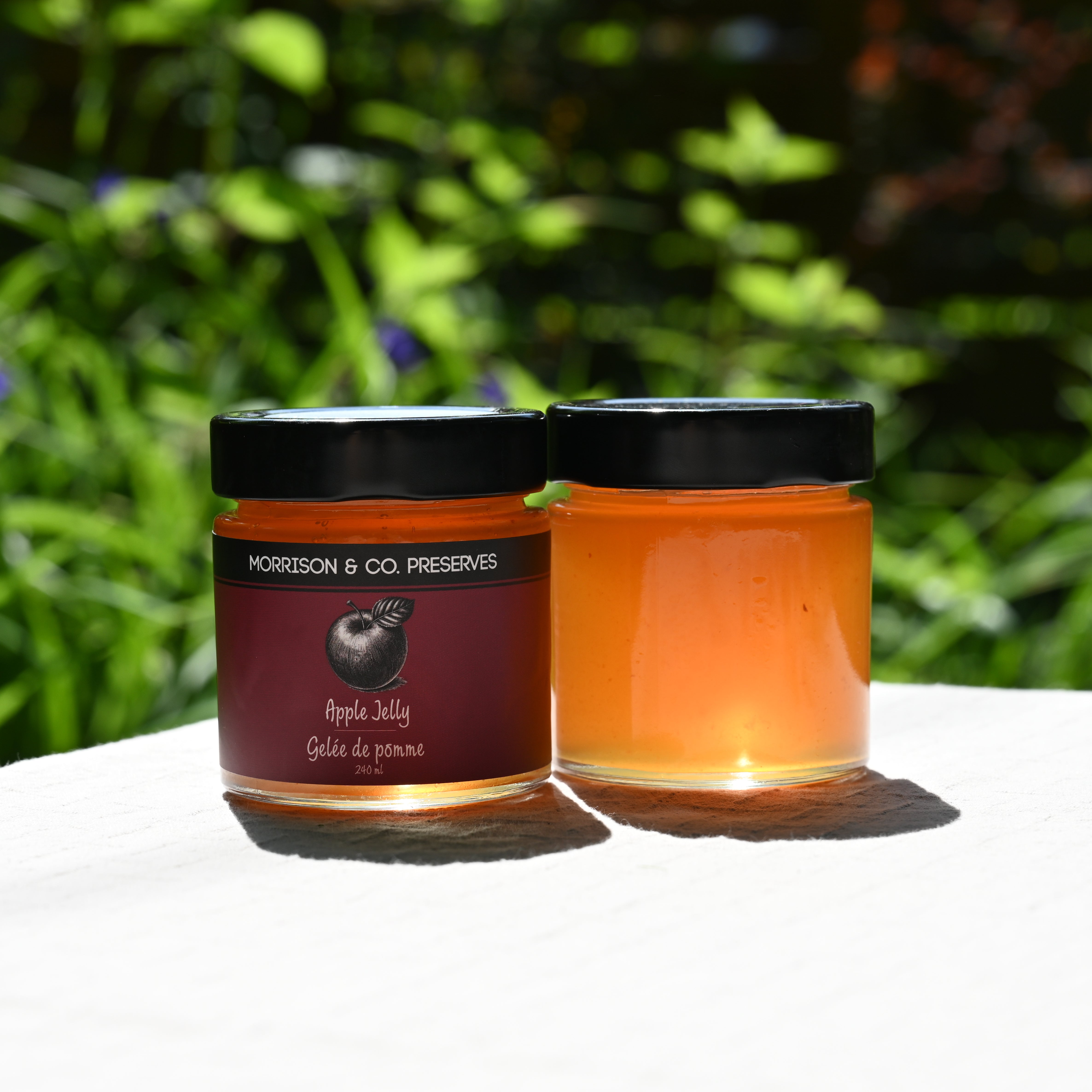 Two jars of apple jelly with a black lid on a white surface, greenery in the background