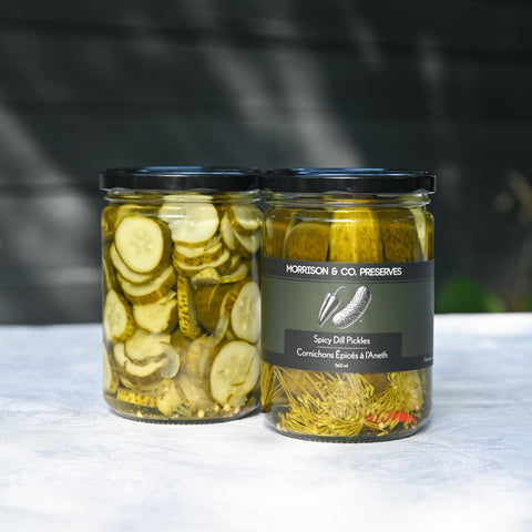 Two glass jars of dill pickles with black lids on a white tablecloth. The left jar contains sliced dill pickles called Ballparks, and the right jar contains spicy spears both with a dark label reading Morrison & Co. Preserves