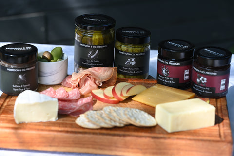 A charcuterie board with  variety of Morrison & Co Preserves. The board has various types of cheese, crackers, cured meats and apples.