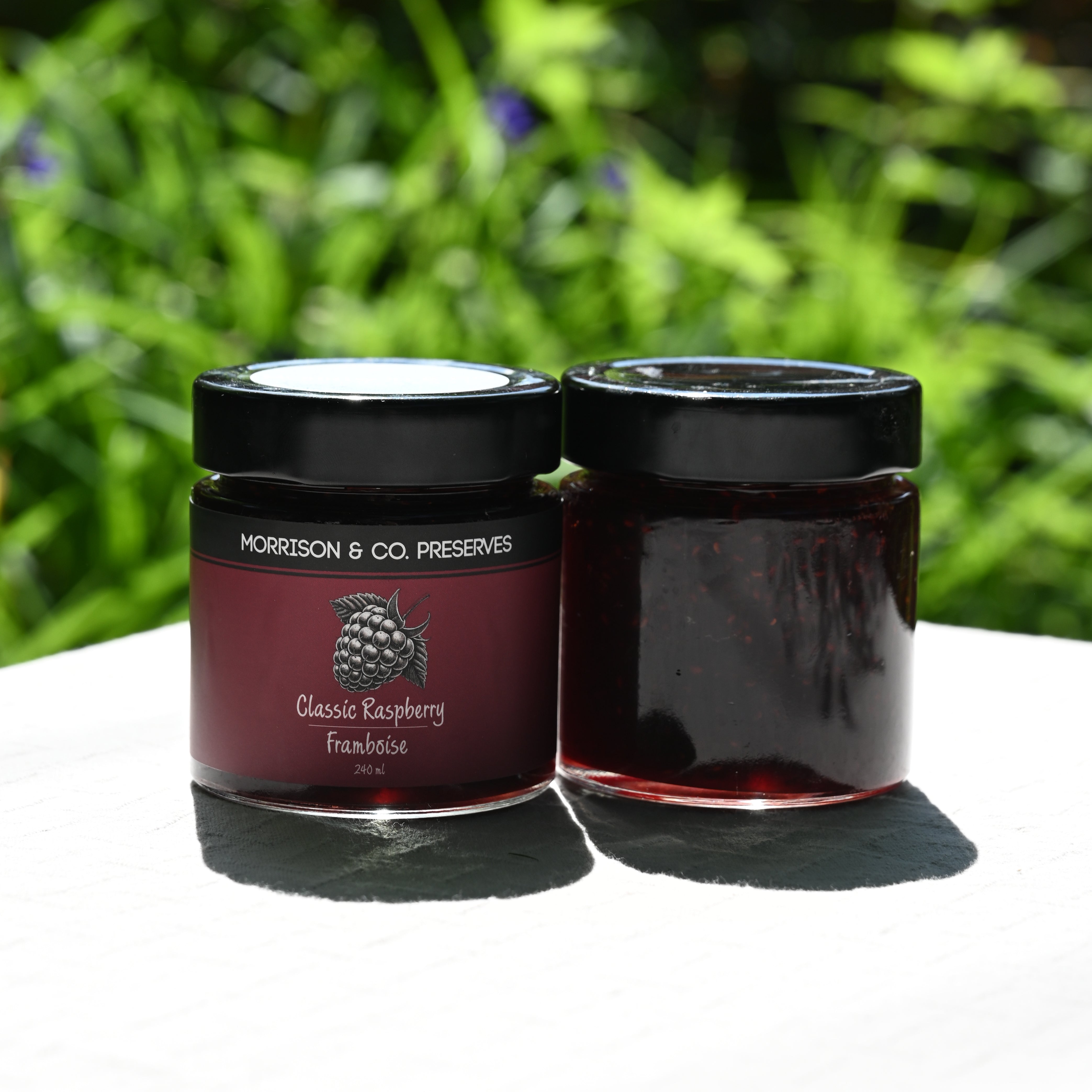 Two jars of Morrison & Co. Preserves Classic Raspberry Jam on a white surface with a green blurred background.