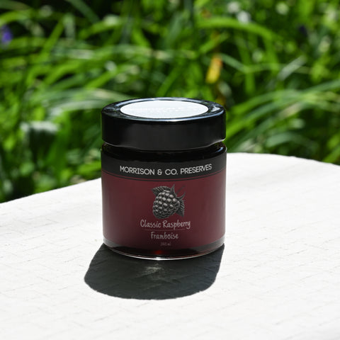 Jar of Morrison & Co. Preserves Classic Raspberry Preserve on a white surface with green grass in the background