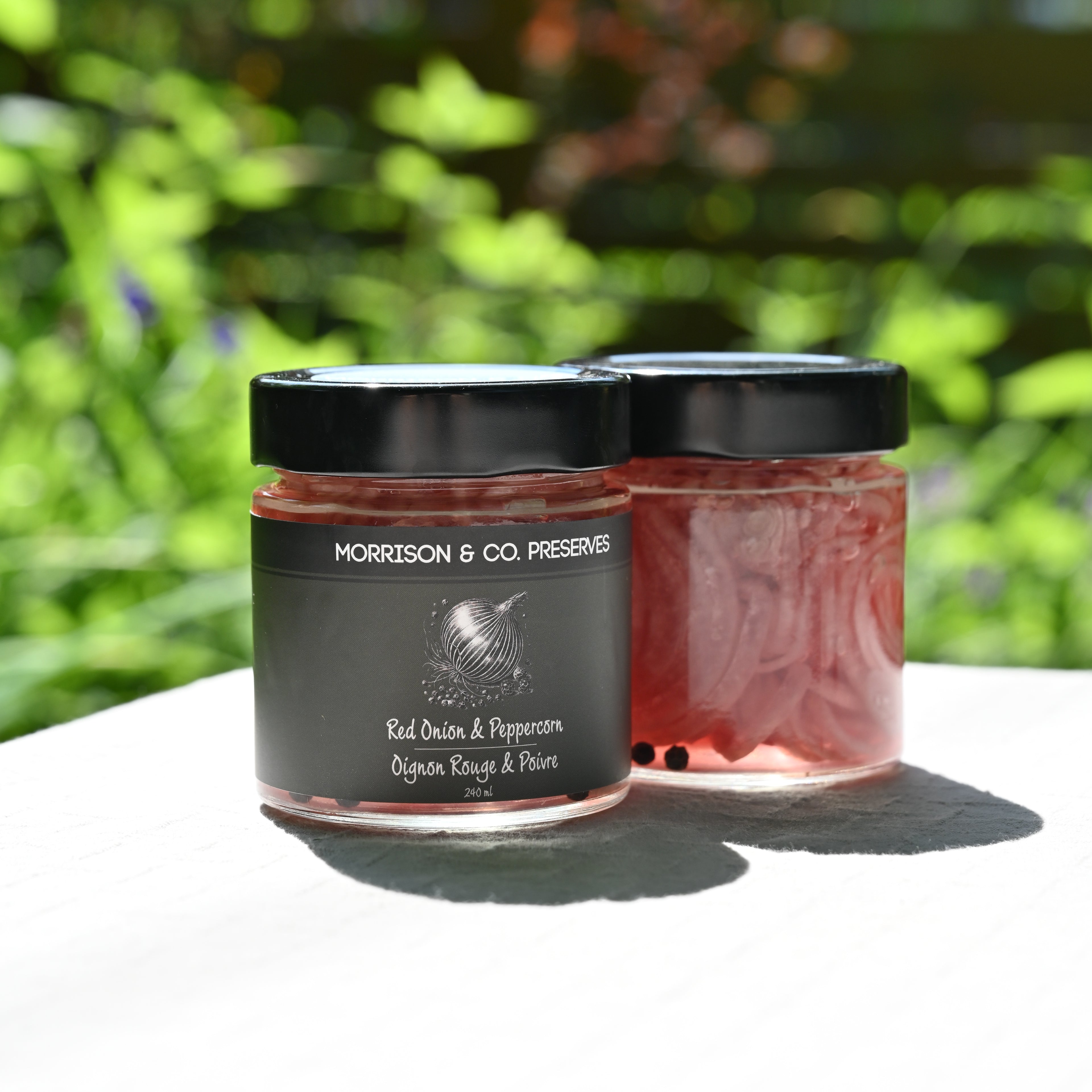 Two jars of Morrison & Co. pickled red onions with a blurred green outdoor background