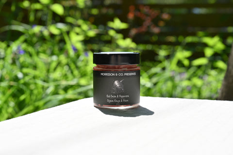 Jar of Morrison & Co. pickled red onion and peppercorns on a white surface with a blurred green outdoor background
