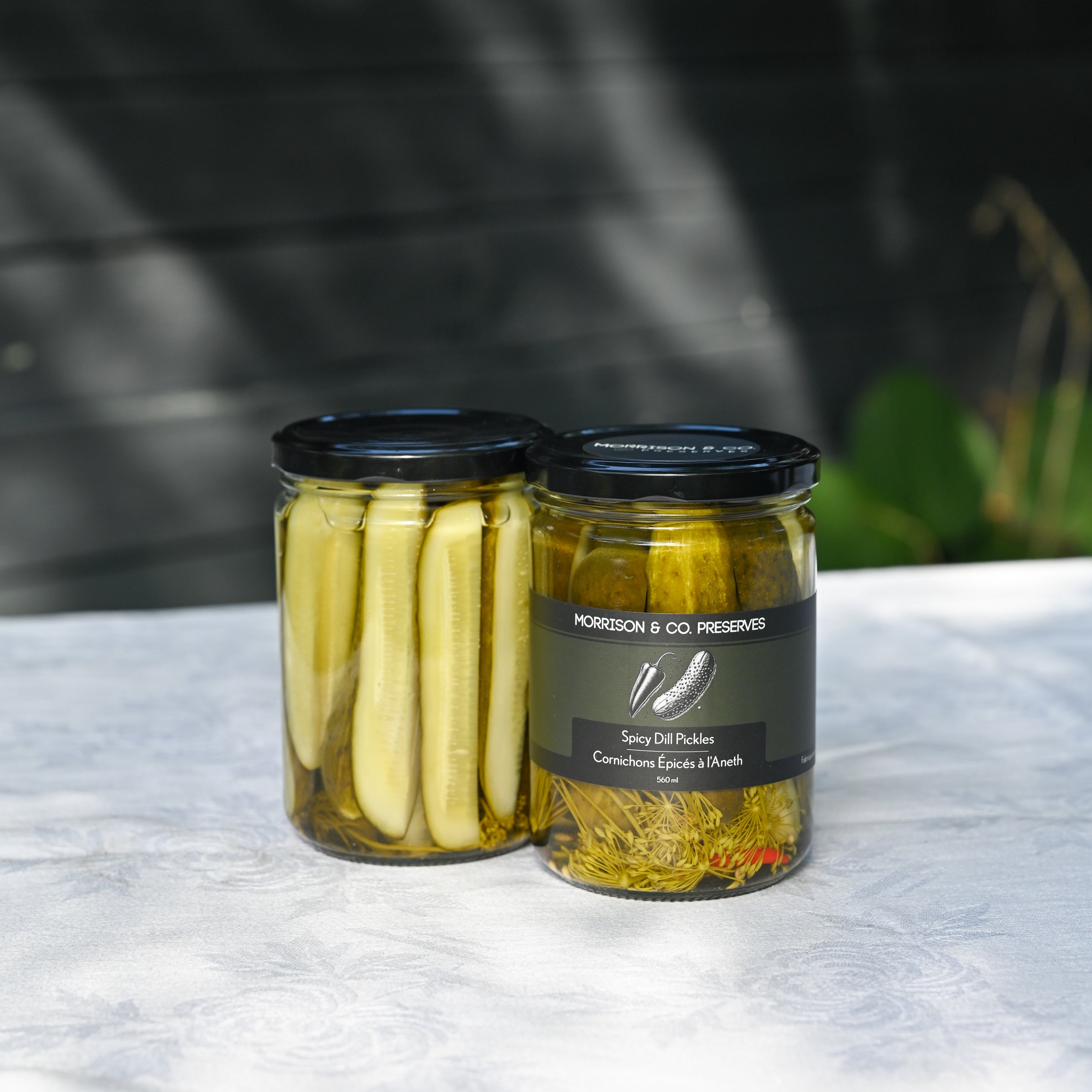 Two glass jars of Morrison & Co Preserves spicy dill pickles on a white textured tablecloth with a dark, blurred outdoor background. 